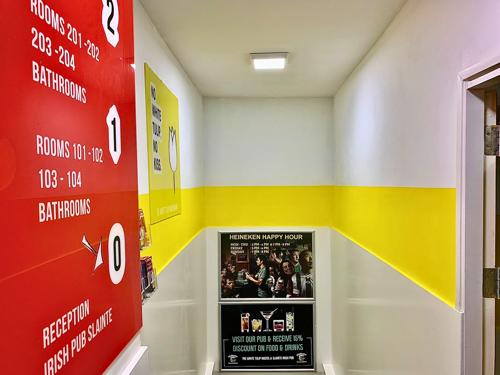 The White Tulip Hostel Amsterdam - 1 A brightly colored corridor with directional signage. To the left, a red wall with white text indicating room numbers and facilities on different floors; ahead, a poster for Heineken Happy Hour; and to the right, a yellow wall with a playful text poster.